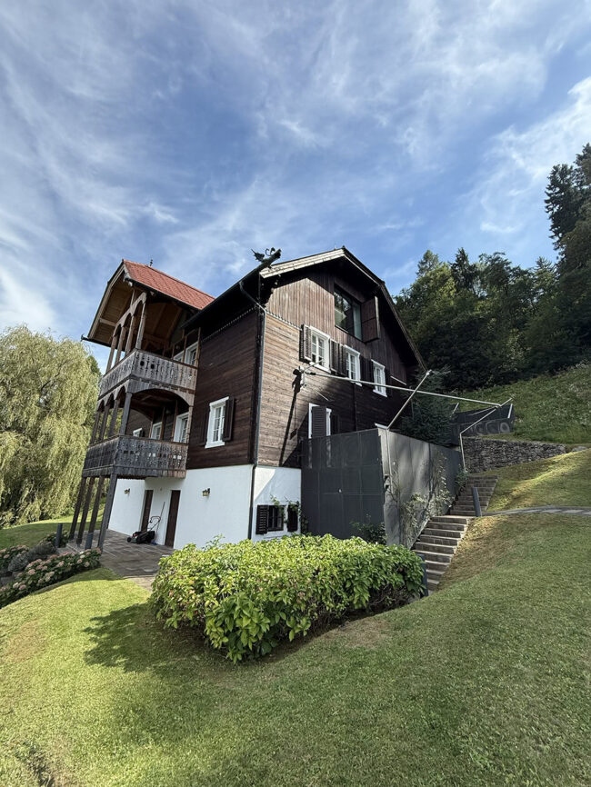 Energieeffizientes Holzhaus mit traditionellem Charme, umgeben von grüner Natur, unter blauem Himmel, attraktive Gartengestaltung und moderne Architektur.