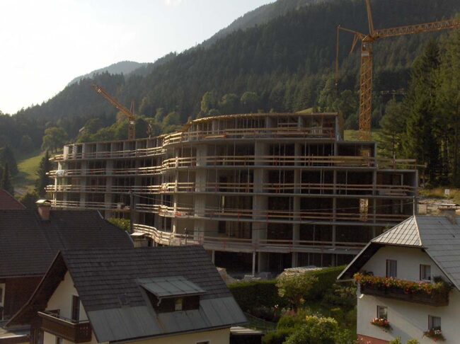 Bau des neuen Mehrfamilienhauses in einer bergigen Landschaft, mit Kränen im Einsatz, im Hintergrund grüne Wälder und Hügel.