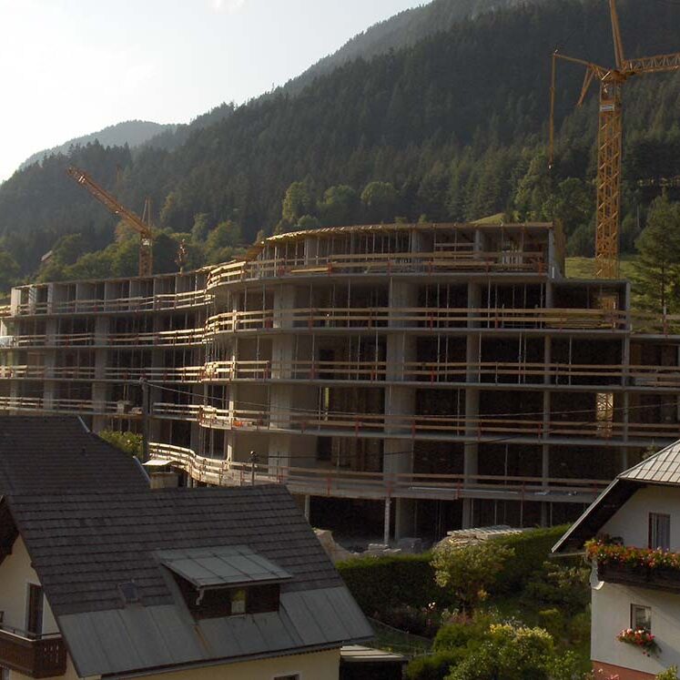 Bau des neuen Mehrfamilienhauses in einer bergigen Landschaft, mit Kränen im Einsatz, im Hintergrund grüne Wälder und Hügel.