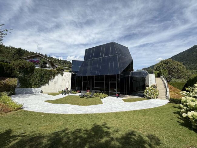 Moderne Architektur eines gläsernen Hauses in alpiner Lage mit Garten und umliegenden Bergen, perfekt für nachhaltiges Bauen und innovative Baukonzepte.