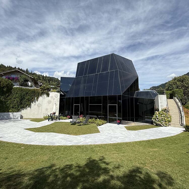 Moderne Architektur eines gläsernen Hauses in alpiner Lage mit Garten und umliegenden Bergen, perfekt für nachhaltiges Bauen und innovative Baukonzepte.