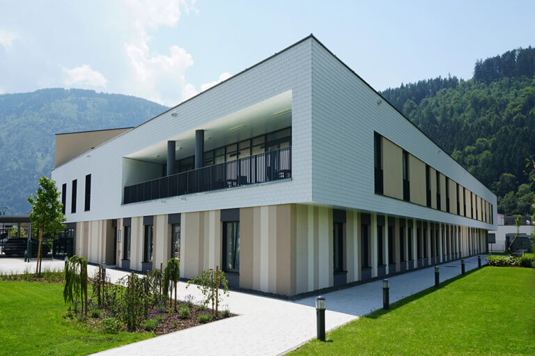 Modernes Bürogebäude in den Bergen mit weißer Fassade, großer Terrasse und modernen Fenstern, umgeben von grünem Rasen und harmonischer Landschaft, ideal für gewerbliche Nutzungen in einer ländlichen Umgebung.