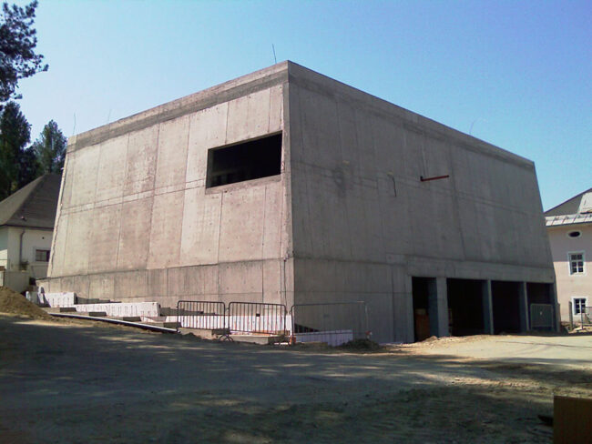 Rohbau eines modernen Gebäudes aus Beton, im Bau befindlich, kaum fertiggestellt, auf einem Baugrundstück in einer Stadt in Österreich, Baustelle mit Sicherheitsbarrieren, klarem Himmel, aktuelle Bauphase.