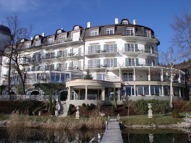 Elegantes historisches Hotel am Wasser mit weißen Fassadenelementen, großzügigen Balkonen und einer Dachterrasse, umgeben von attraktiver Gartenanlage und Wasserpromenade, ideal für Wellness- und Erholungssuchende.