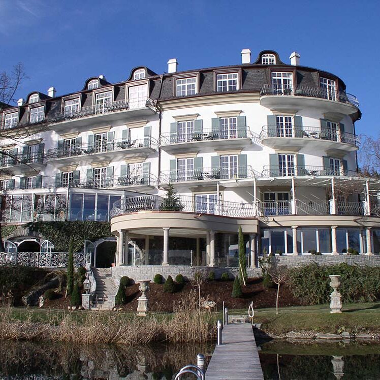 Elegantes historisches Hotel am Wasser mit weißen Fassadenelementen, großzügigen Balkonen und einer Dachterrasse, umgeben von attraktiver Gartenanlage und Wasserpromenade, ideal für Wellness- und Erholungssuchende.