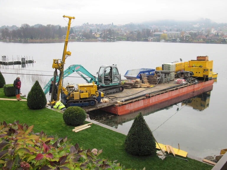 Betonboden wird auf Fluss mit Baumaschinen und Bagger repariert, Bauarbeiten am Wasser, Baustelle am Ufer mit Boot und Landschaft im Hintergrund - Infrastruktur, Wasserbau, Sanierung.