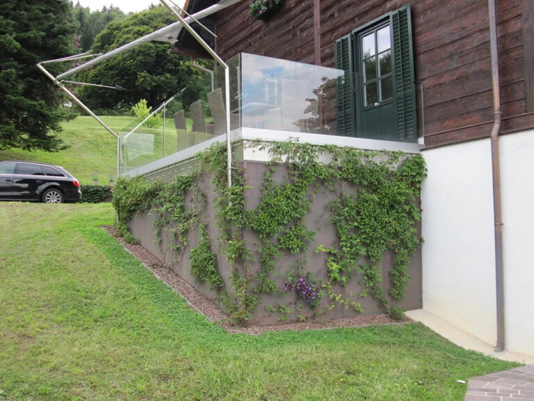 Gartenveranda mit Glasgeländer, grüne Pflanzen und Blick auf die grüne Landschaft, rustikale Holzfassade, ideal für Erholung und Naturverbundenheit.