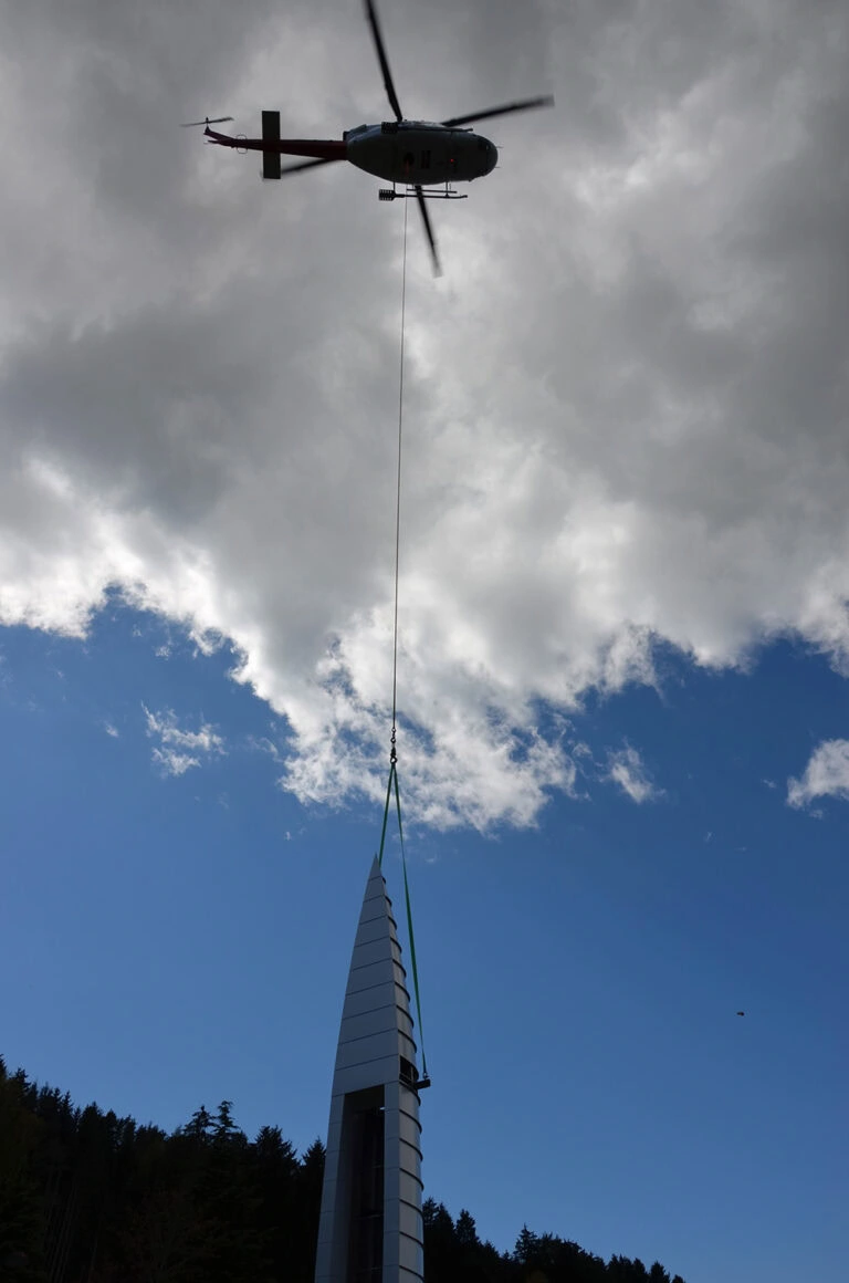 Helikopter im Einsatz bei Bauarbeiten an einem modernen Gipfelkreuz auf einem hohen Berg, symbolisch für innovative Bauprojekte und Handwerkskunst.