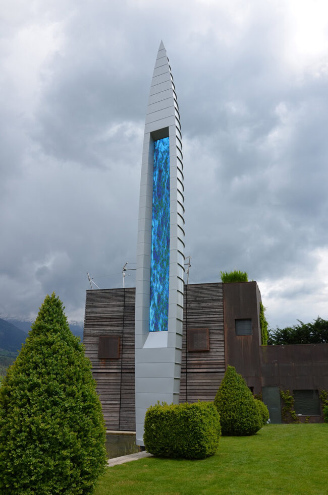 Moderner, langer LED-Bildschirm auf einer Gebäudefassade, umgeben von grünen Sträuchern und Bergen, bei bedecktem Himmel, als architektonisches Highlight und Blickfang in urbaner Umgebung.