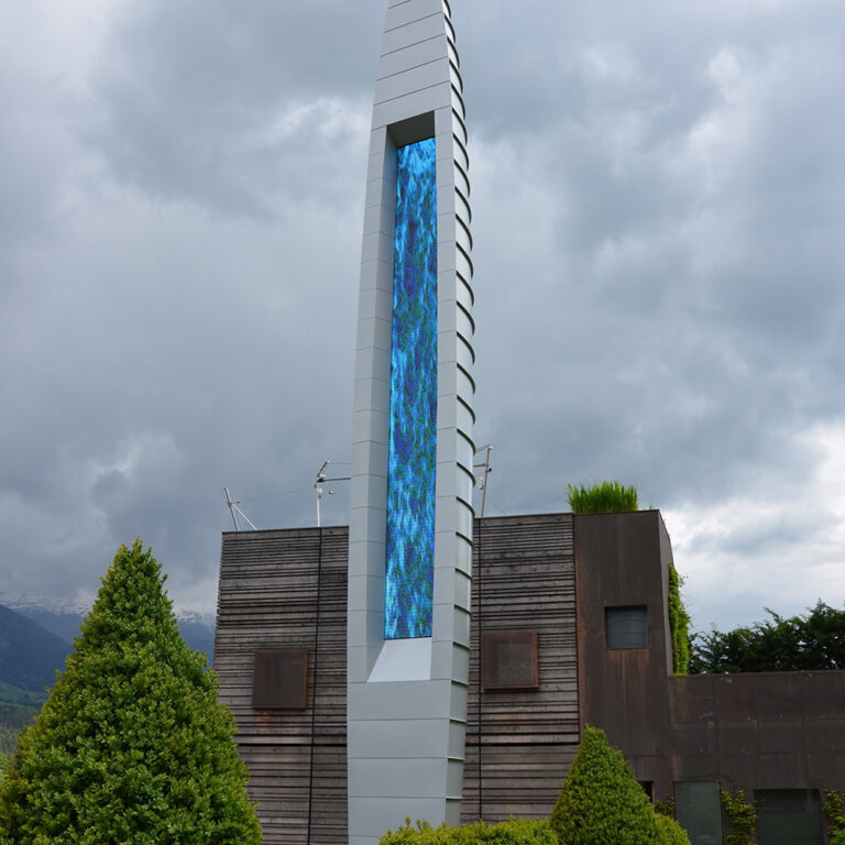 Moderner, langer LED-Bildschirm auf einer Gebäudefassade, umgeben von grünen Sträuchern und Bergen, bei bedecktem Himmel, als architektonisches Highlight und Blickfang in urbaner Umgebung.