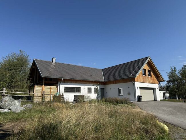 Modernes Landhaus mit Holzfassade und großen Fenstern, umgeben von Grünfläche, ideal für erholsames Wohnen im Grünen.